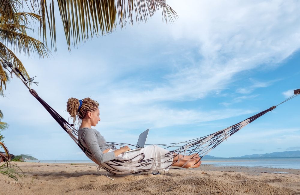 Chica trabaja a distancia con su portátil en una hamaca suspendida sobre la arena de la playa