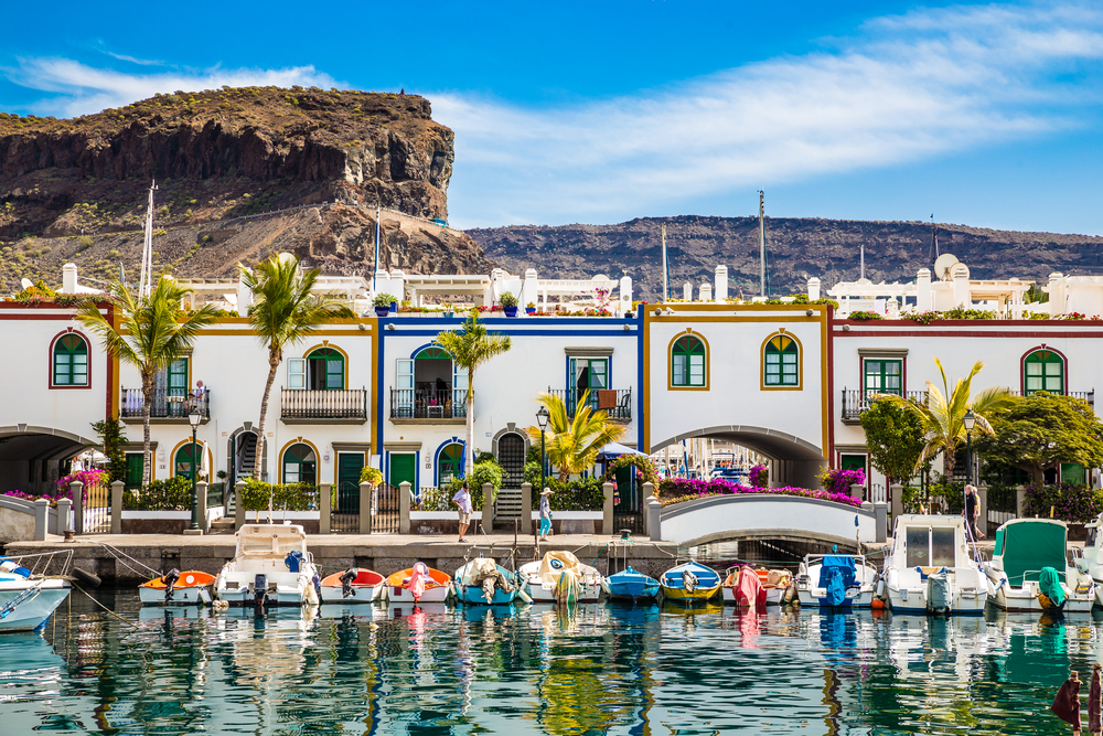 Edificios De Colorido Tradicional Con Barcos En Frente Y Montaña En El Fondo - Puerto De Mogán, Gran Canaria