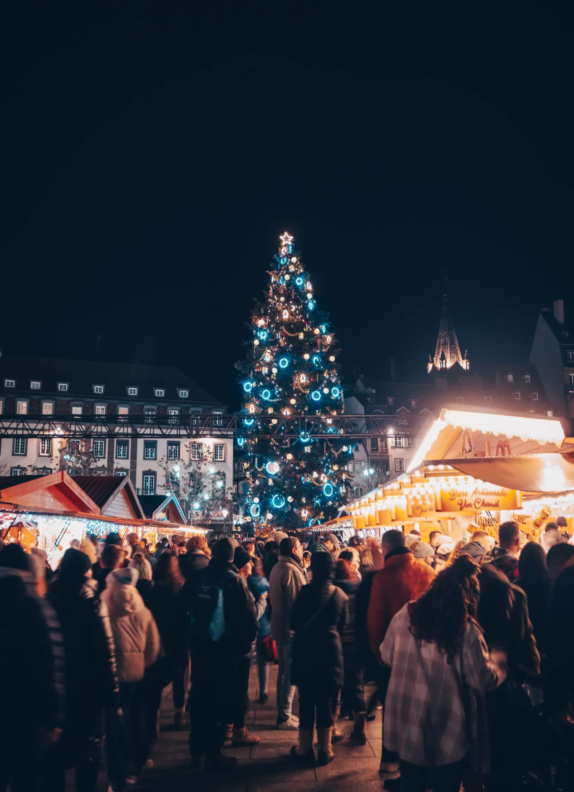Un concurrido mercado navideño de Estrasburgo de noche, con un imponente árbol de Navidad iluminado con luces azules y blancas, rodeado de puestos tradicionales de madera y visitantes abrigados.